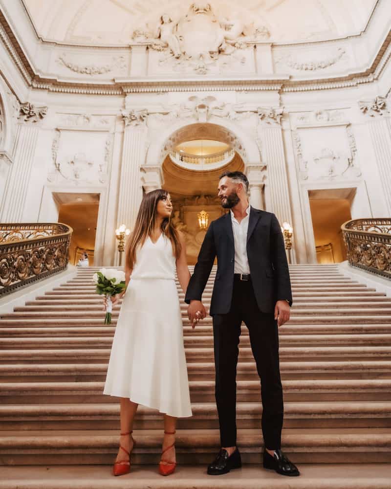 a couple posing on the grand staircase at city hall - bride has a uniquely cut dress and red shoes
